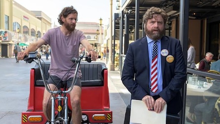 Zach Galifianakis wearing American flag tie with buttons on suit with man riding bike carriage in FX's Baskets