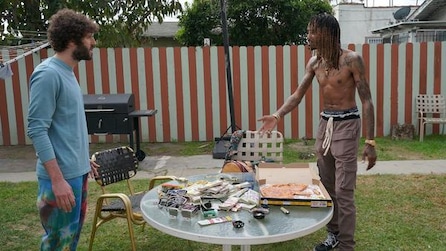 Dave talking with GaTa in backyard with GaTa pointing to table with cash and pizza in FX's Dave