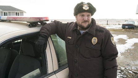 Man with beard in police winter uniform leaning against police car with melted snow in background in FX's Fargo Year Three