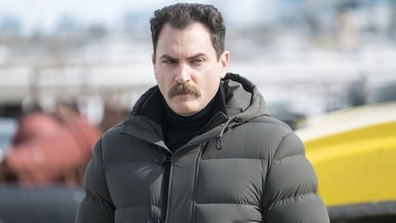 Man with mustache in black puffy jacket walking outside sunny day with yellow car in background in FX's Fargo Year Three