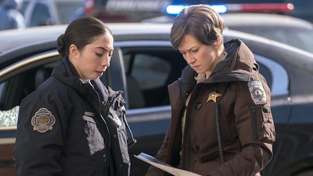 Carrie Coon as Gloria looking at report with female officer in front of black car and police cars in FX's Fargo Year Three