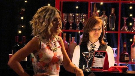 Blonde woman in colorful dress leaning on bar with bartender behind looking down in FX's Legit