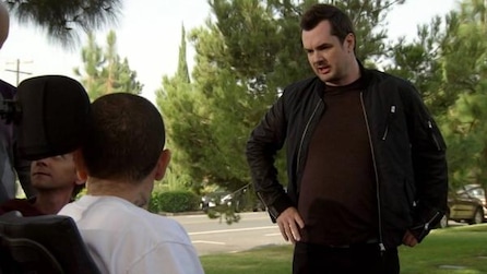 Jim Jefferies looking down at man with neck tattoo in white shirt outside by trees in FX's Legit