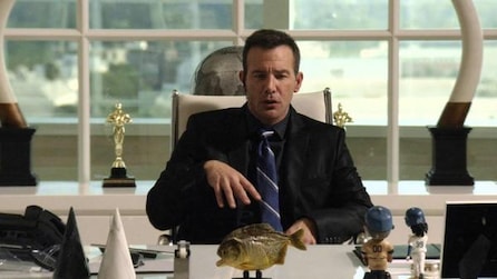 Man in suit talking behind white desk with fish desk statue and Oscar award in background in FX's Legit