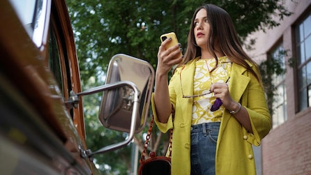 Woman wearing a yellow coat holding a phone and standing next to a car