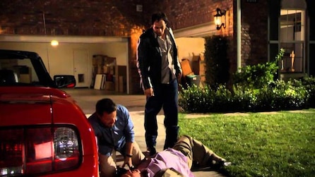 Man laying on the floor next to a car while two other men hover over him