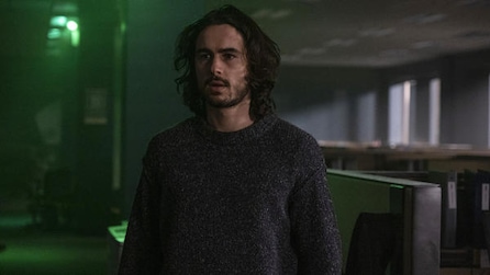 Man with long black hair wearing a sweater standing in a dark room