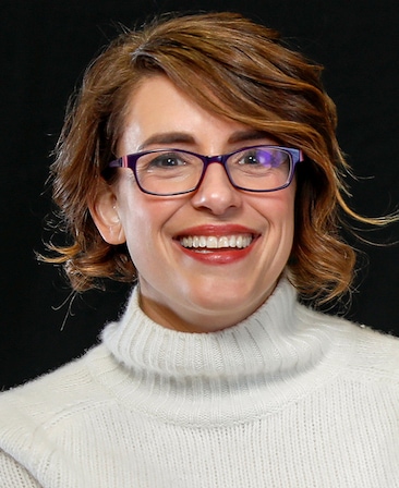 Anna Boden headshot wearing a white sweater and black glasses