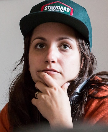 Hannah Fidell Headshot wearing a green hat and placing her hand on her chin