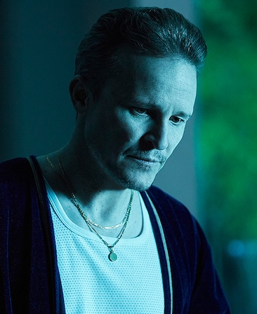Damon Herriman headshot wearing gold necklaces, a white shirt and black jacket