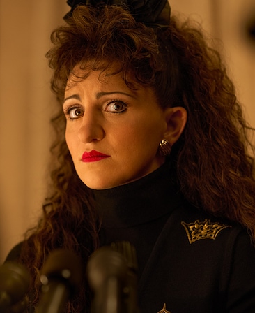 Headshot of Annaleigh Ashford as Paula Jones in black coat and bow in front of microphones from FX's American Crime Story