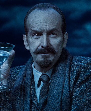 Headshot of Denis O'Hare with mustache in grey suit holding martini glass on cloudy night from FX's AHS Double Feature