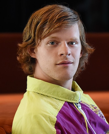 Lucas Hedges headshot wearing a yellow and purple jacket 