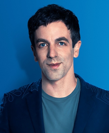 B.J. Novak headshot wearing a teal shirt, navy jacket and standing in front of a blue wall