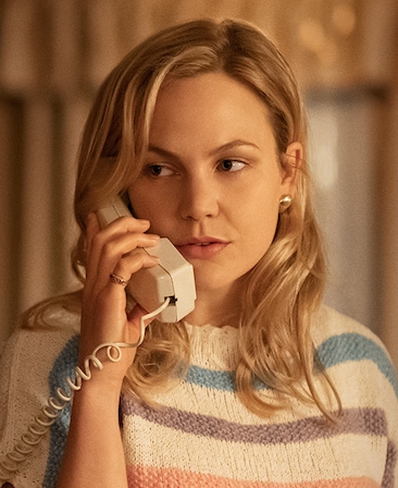 Adelaide Clemens headshot wearing a white blue and purple striped sweater and holding a phone with cord to her ear