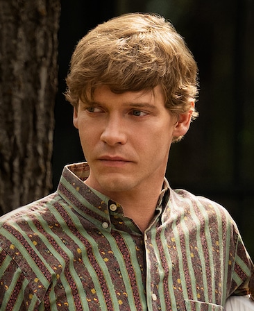 Billy Howle headsot wearing a brown and green striped shirt standing by a tree