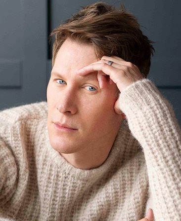 Dustin Lance Black headshot wearing a knit cream sweater with hand on his head