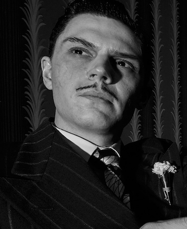 Black and white headshot of Evan Peters with thin lined and short mustache in black striped suit with white boutonniere