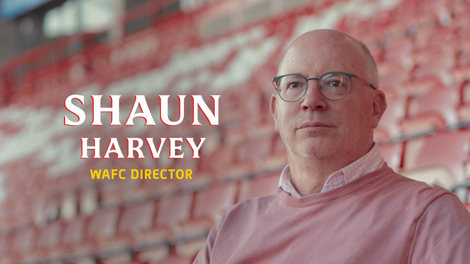 Shaun Harvey headshot wearing a pink sweater with a pink checkered button up underneath, sitting in the stands and his name.