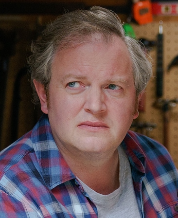 Miles Jupp headshot wearing a white shirt and red and blue plaid top sitting inside