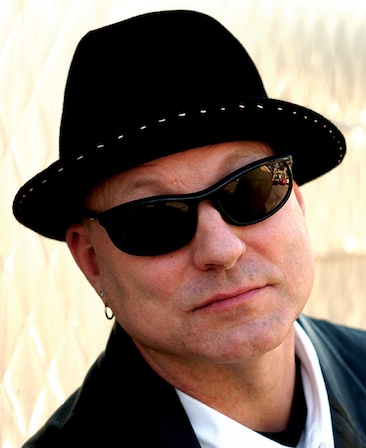 Charles H Eglee headshot wearing a black fedora with white stitching and black sunglasses, with an earring in his ear