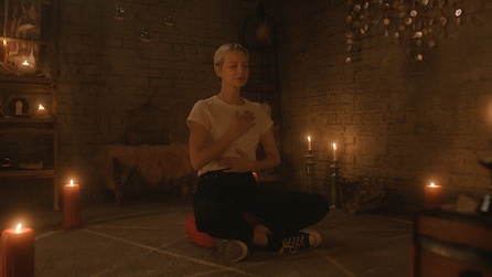 Woman with short blonde hair sitting cross legged in the middle of a pentagram with candles around her