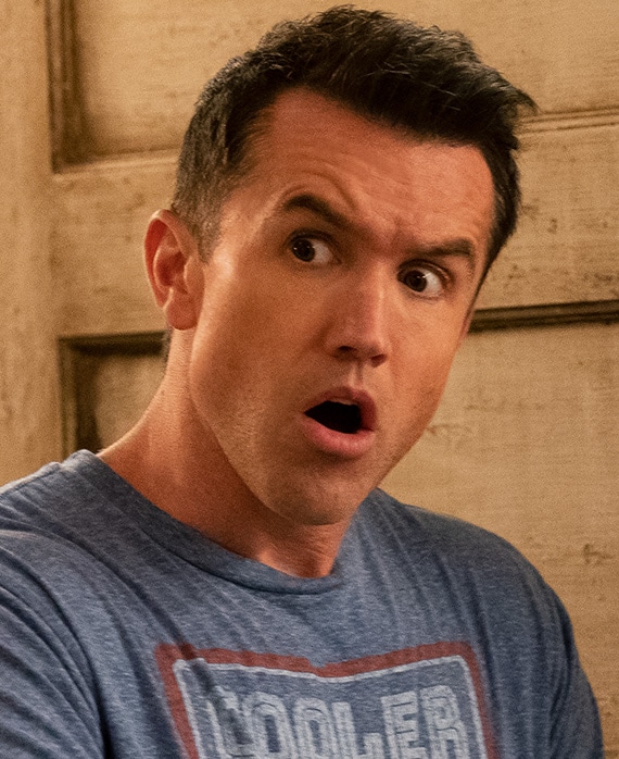 Rob McElhenney headshot wearing a gray shirt that reads "cooler"an standing in front of a wood background