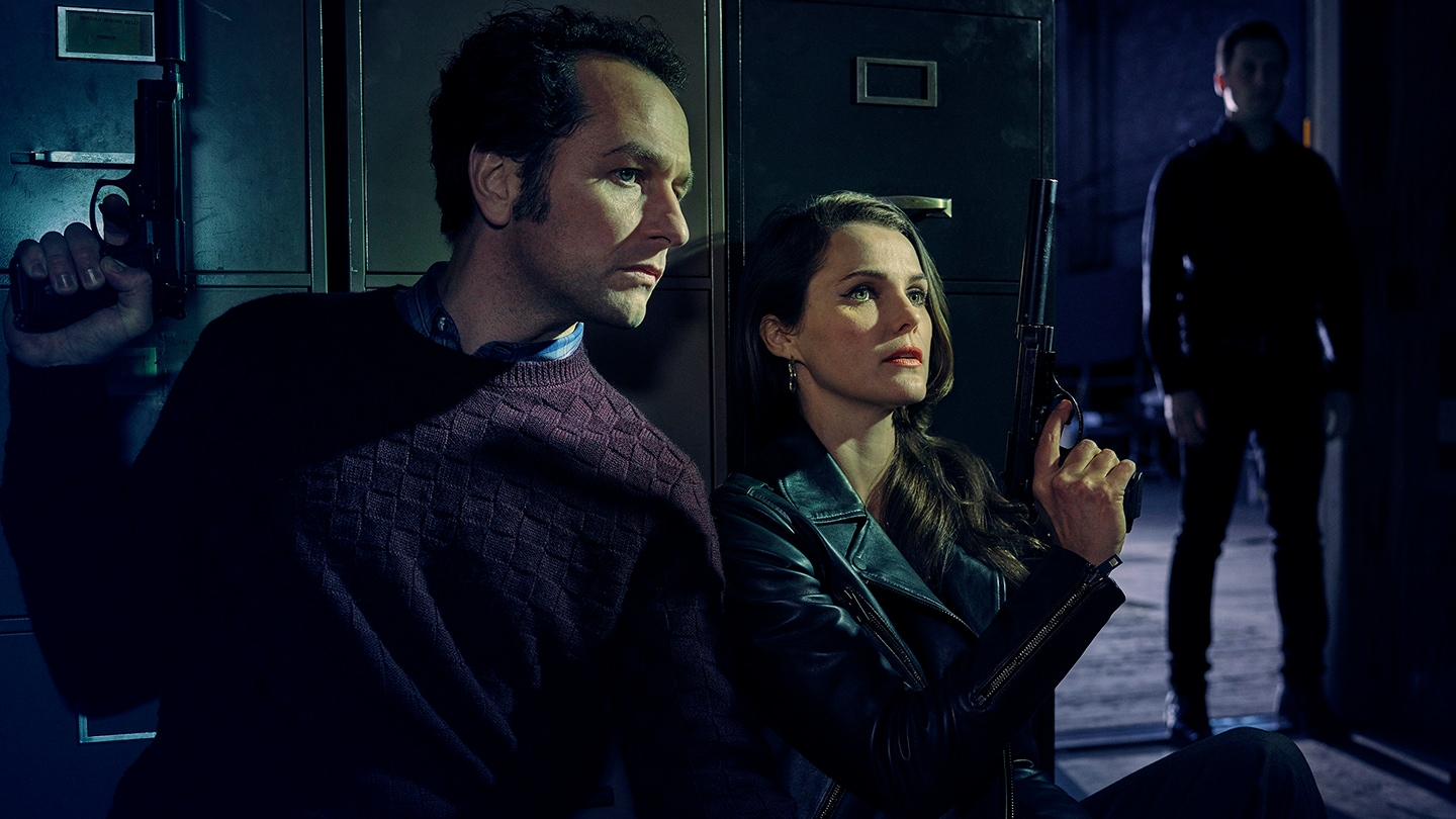 Keri Russell as Elizabeth Jennings, Matthew Rhys as Philip Jennings sitting and hiding for a shadowed figure.