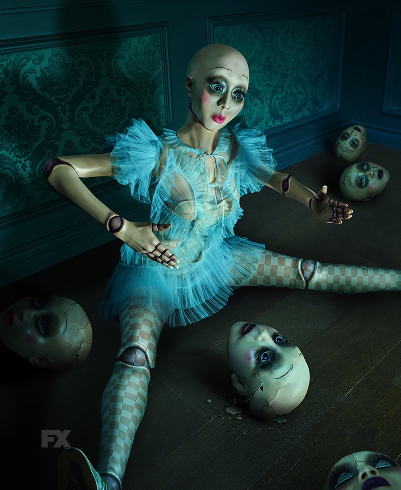 Doll on floor wearing blue dress and checkered tights surrounded by doll heads from FX's American Horror Stories