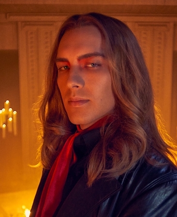 Cody Fern headshot in black suit with red scarf in front of bright candles from AHS Apocalypse