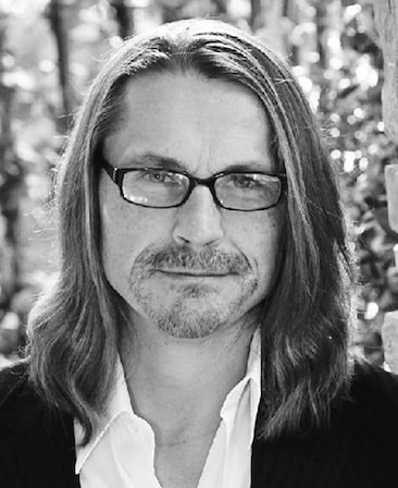 Kurt Sutter headshot wearing a white shirt with a black jacket and standing outside