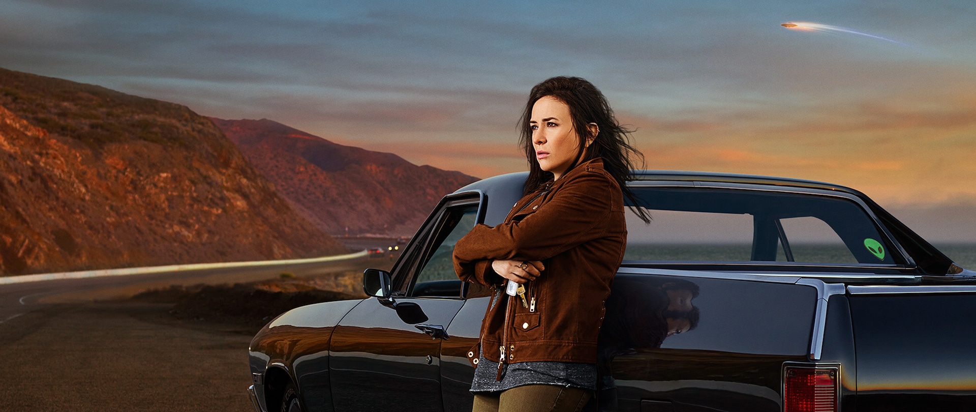 Sam from Better Things in brown jacket leaning against black impala with sunset and mountains in background