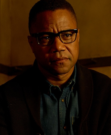 Cuba Gooding Jr. in blue denim button down with black cardigan and wearing glasses in dark white room with half shadowed face