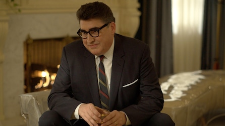 Alfred Molina as Robert Aldrich sitting down on chair holding drink with fireplace in background in Feud FX show