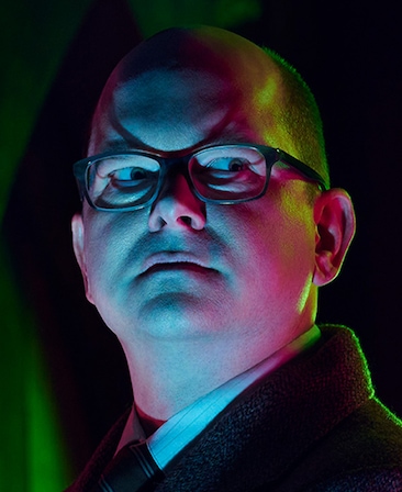 Headshot of Mark Proksch as Colin Robinson wearing glasses in a suit and tie looking off to his side.
