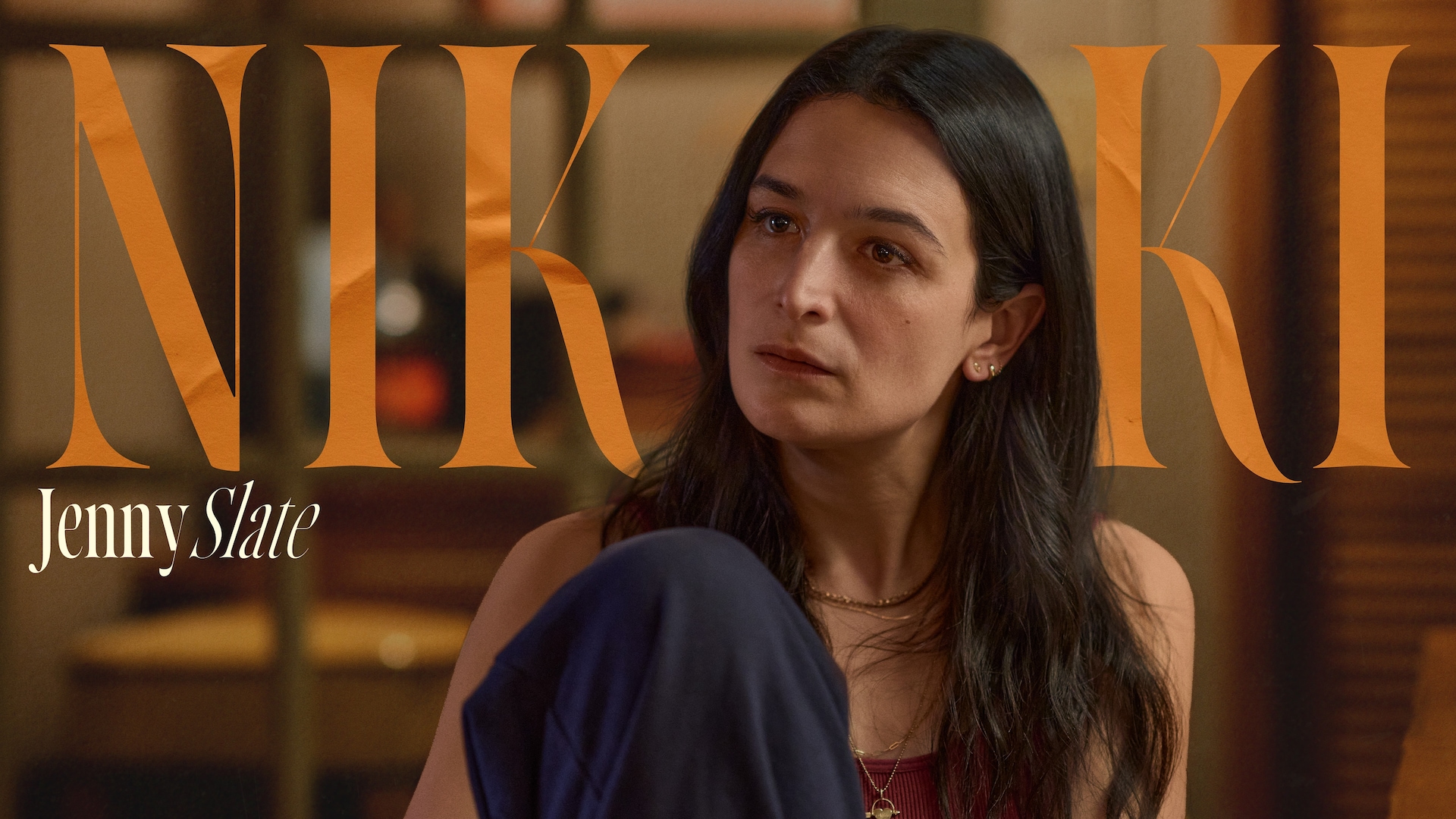 Jenny Slate as Nikki Boyer in FX's Dying For Sex, wearing navy blue pants sitting with her knee to her chest.