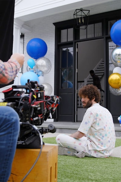Dave Burd sitting on grass with blue and silver balloons in front of a camera filming an episode of Dave FX show