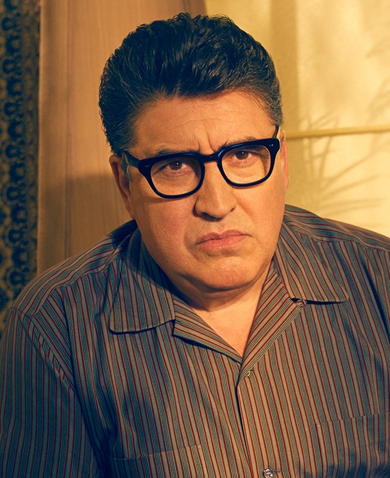 Alfred Molina as Robert Aldrich wearing green striped shirt and black glasses in front of yellow window drapes in Feud FX show