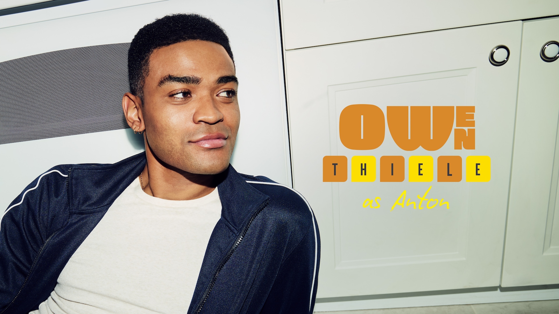 Owen Thiele as Anton in FX's Adults, wearing a white shirt, navy blue stripped jacket, leaning against an oven.