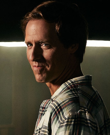 Nat Faxon headshot wearing a plaid shirt standing in front of a light