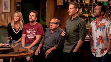 Cast of It's Always Sunny sitting against bar top looking at obscured figure with bag on table in FX's It's Always Sunny