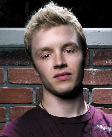 Noel Fisher headshot a burgundy shirt and standing in front of a brick wall