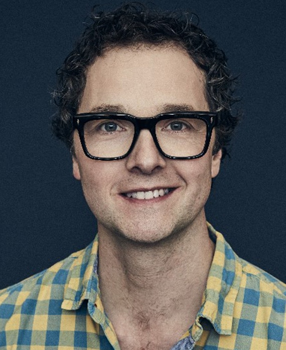 Chris Addison headshot in light yellow and blue checkered flannel with dark navy background
