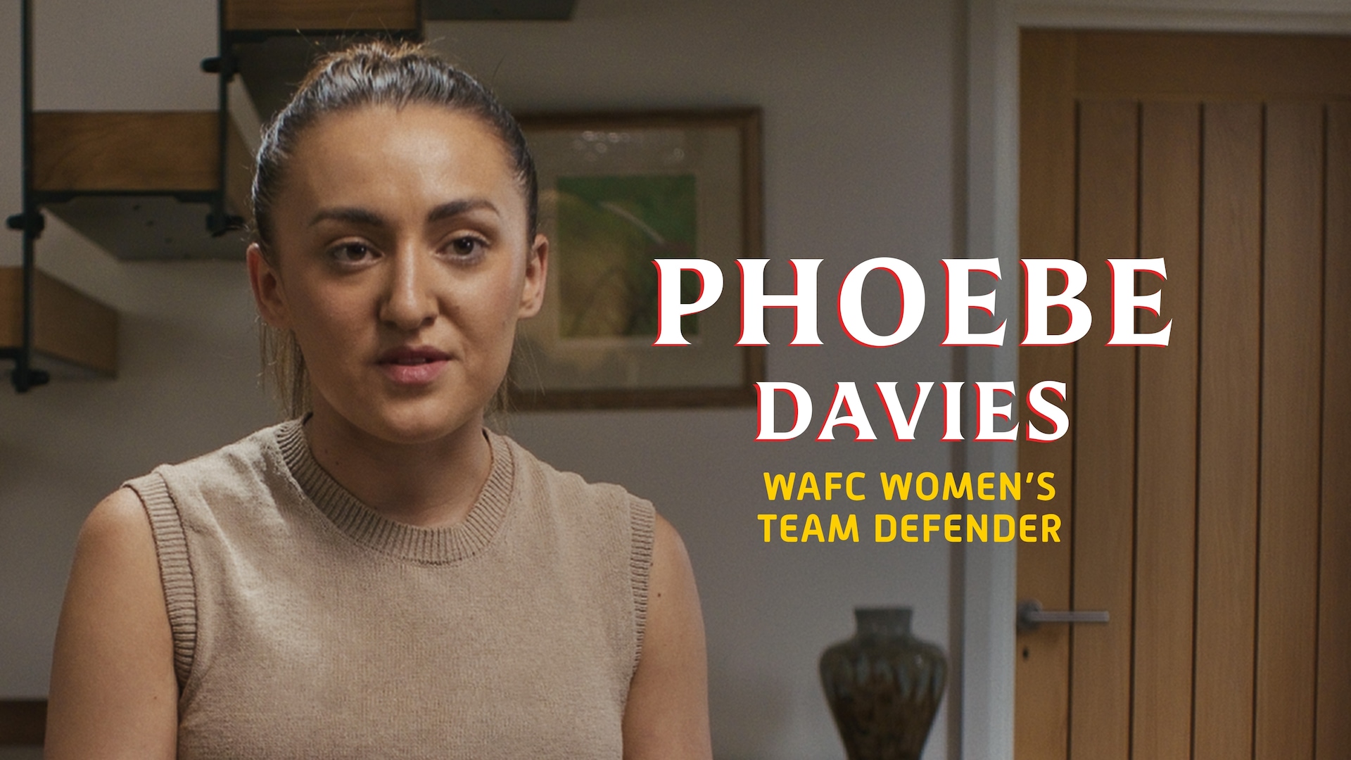 Phoebe Davies headshot, wearing a brown sweater with her hair tied back, sitting in a room with her name in large text.