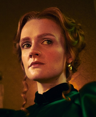 Gayle Rankin headshot in dark green velvet high neck dress, with hair up in bun with ringlets hanging down either side