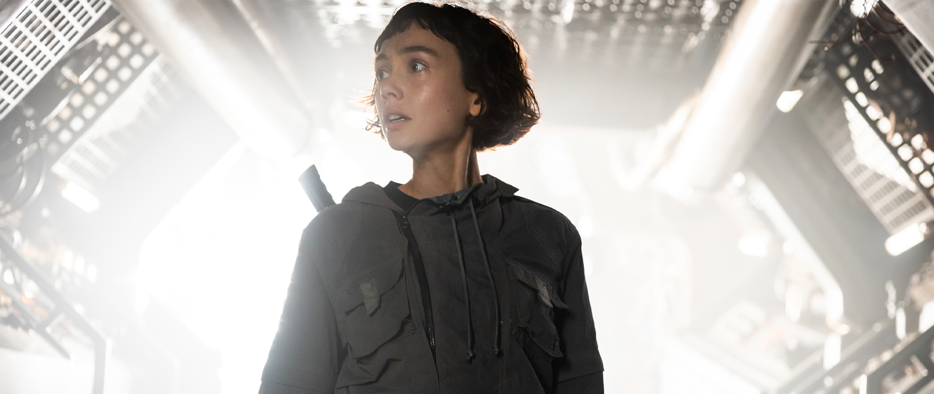 Woman with short brown hair wearing a grey utility suit and looking off in the distance with a white glow behind.