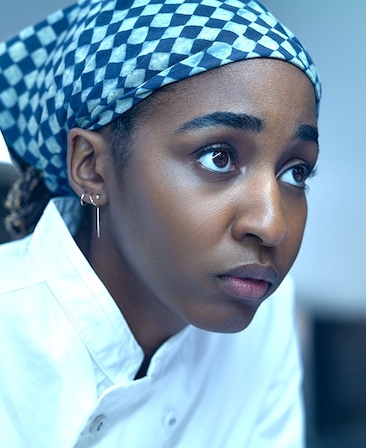 Ayo Edebiri headshot wearing a white chef jacket and a blue and white checkered bandana.