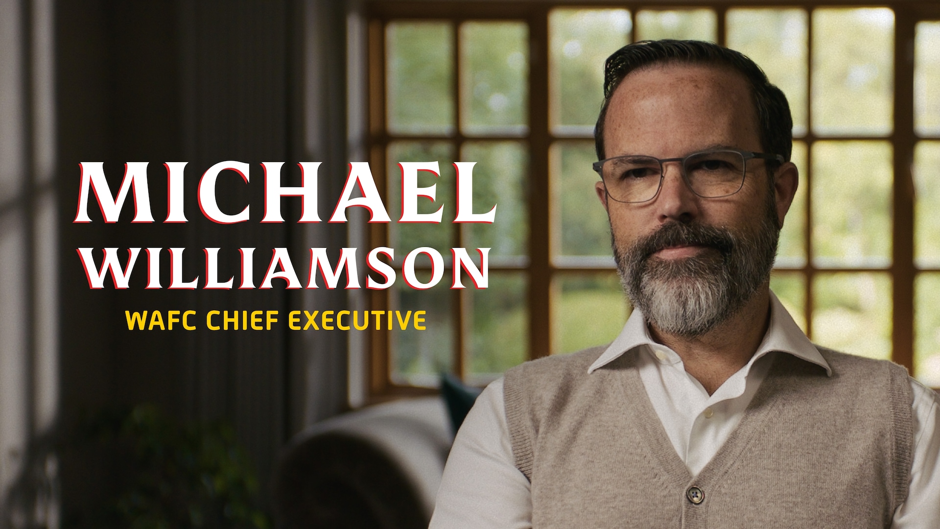 Michael Williamson headshot wearing a brown sweater over a white button up, in beige rimmed glasses, in front of a window.