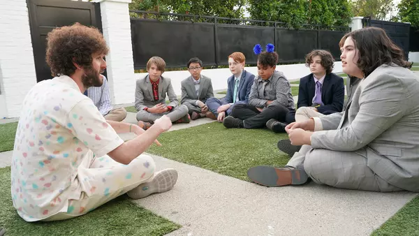 Dave in pastel dotted shirt sitting on grass and concrete with young boys in circle in FX's Dave