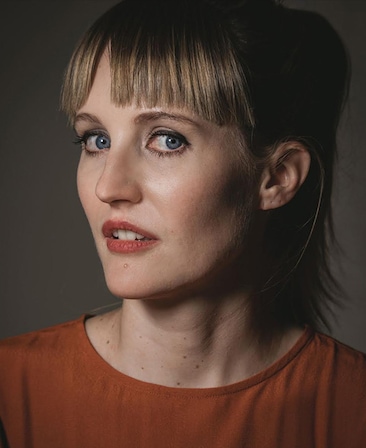 Headshot of Shannon Murphy wearing an orange blouse with her hair up in a pony tail, standing in front of a beige background.